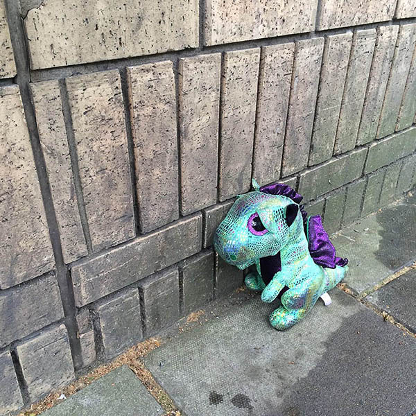Abandoned, unwanted, unloved, cuddly toy - Sad looking, iridescent dinosaur on the Pavement, leaning against a brick building
