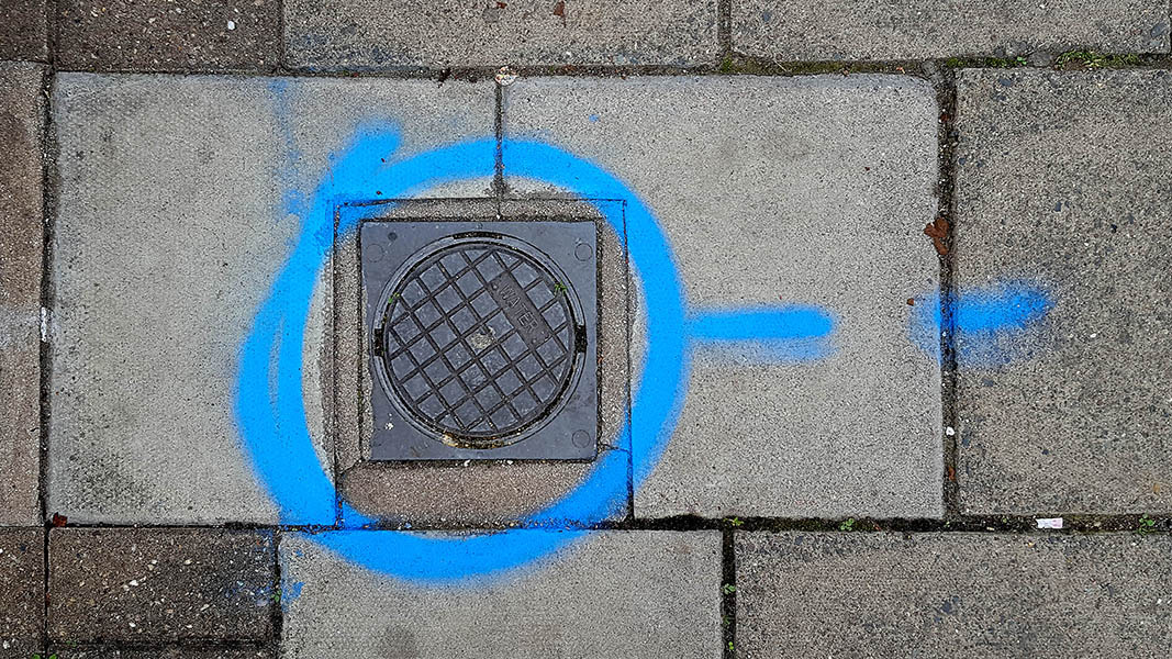 Pavement markings - spray painted squiggles on paving stones - Blue circle surrounding small water access panel with a broken line to the right